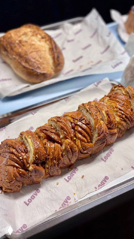 Ассортимент пекарни Loave на подносе: несколько буханок и завитая карамельно-ореховая булка на бумаге с логотипом кафе, интерьер фона.