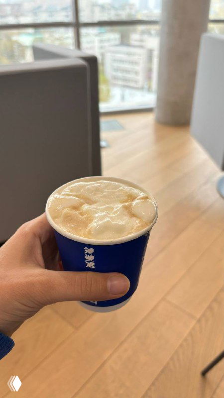 Рука держит бумажный стакан с кофе и плотной творожно-сырной пенкой на фоне окна с видом на город и интерьер кофейни.