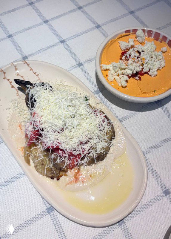 Запечённый баклажан с фетой, томатами и несколькими соусами, посыпанный тёртым сыром, кадр крупным планом на тарелке.