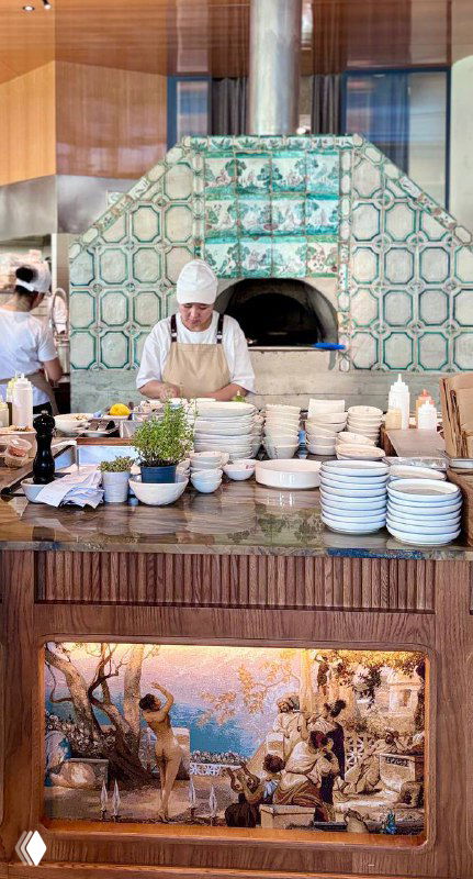 Открытая кухня и печь с орнаментальной плиткой, повар за работой и стойка с тарелками — видно кулинарный антураж заведения.