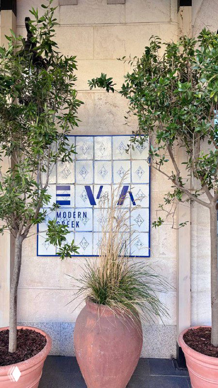 Фасад ресторана с плиточной вывеской "EVA" и горшками с растениями у входа, приветливый городской вход рядом с метро Киевская.