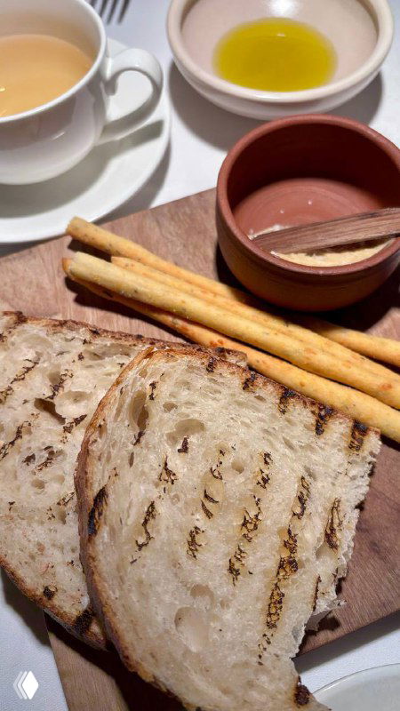 Тёплые ломтики хлеба и хрустящие хлебные палочки на деревянной доске, рядом чашки с маслом и соусами — деталь подачи и сервировки в Luigi`s garden, Москва.