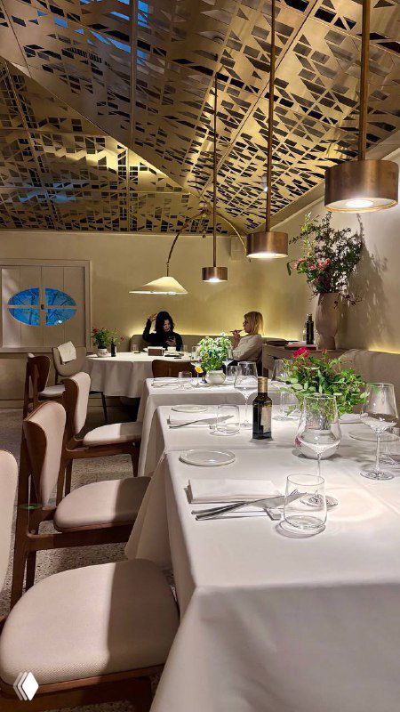 Зал ресторана с гостями за столиками, белые скатерти, зелёные растения и приглушённое освещение — общий вид атмосферы и обстановки.