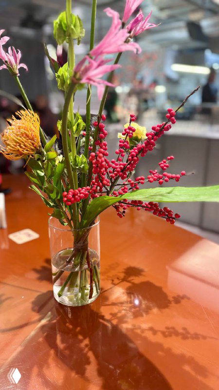 Ваза с высоким букетом цветов на столе внутри заведения; на фоне видна рабочая зона кухни и интерьер кафе с людьми.