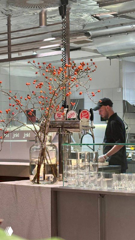 Барная зона и повар за линией раздачи в современном интерьере заведения; на переднем плане высокие вазы с ягодными ветками.