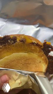 Разрезанный бургер в руке, видно сочную котлету и расплавленный сыр; фольга и упаковка указывают на формат to-go или подачу.