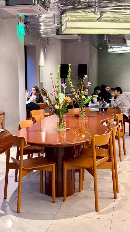 Широкий план зала кафе с длинным общим столом, сидящими гостями и вазами с цветами — уютная атмосферная сцена.