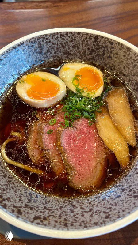 Миска с бульоном и лапшой, два маринованных яйца, ломтики говядины и зелёный лук — тёплое блюдо на столе