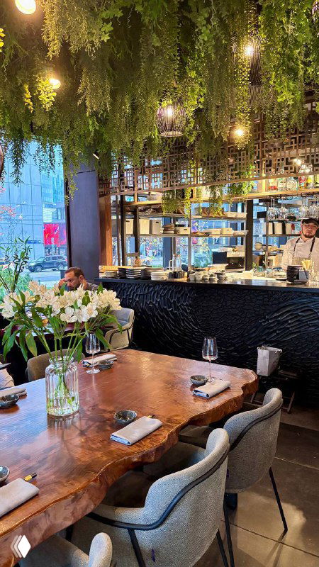 Интерьер ресторана Bluefin: деревянный стол с сервировкой, подвесные зелёные растения и барная зона у больших окон
