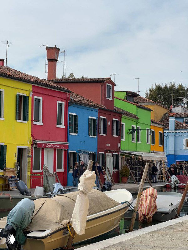 Яркие разноцветные фасады домов острова Бурано вдоль канала, лодки у причала и люди на набережной — типичная сцена венецианского острова.