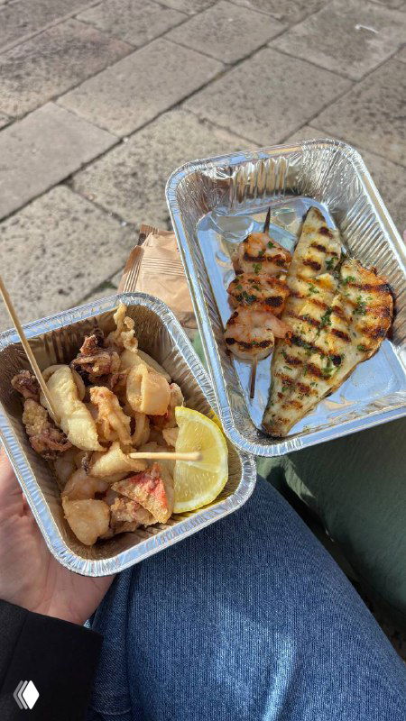 Уличная еда во фольге: чикетти и кусочек рыбы в алюминиевых контейнерах, видно на коленях автора у канала во время быстрой перекуски.