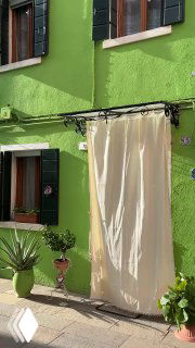 Крупный план зелёного фасада дома с тёмно-зелёными ставнями и белой занавеской, мощёная улочка — бытовая сцена островной Венеции.