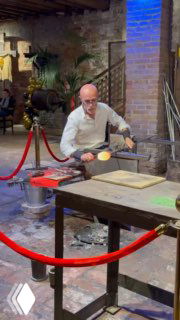 Мастер муранского стеклодува в мастерской за рабочим столом демонстрирует технику выдувания стекла, рядом инструменты и нагретая печь.