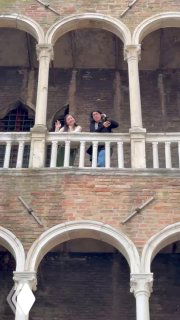 Группа людей выглядывает с верхней галереи арочного исторического здания в Венеции, видны каменные колонны и фасад с окнами.
