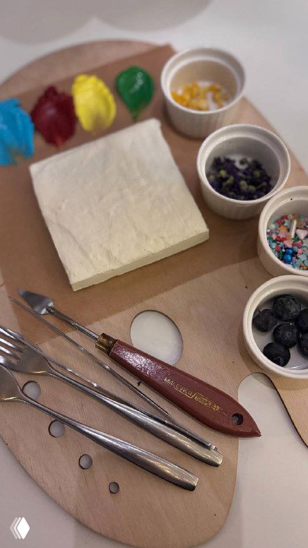 Деревянная палитра с квадратным бисквитом, мисочками со съедобными красками, ягодами и инструментами для росписи торта на столе.