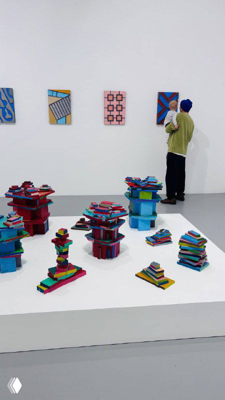 Инсталляция и коллекция маленьких разноцветных геометрических скульптур на белом подиуме в светлом выставочном зале Мультимедиа арт музея Москвы, люди рядом.