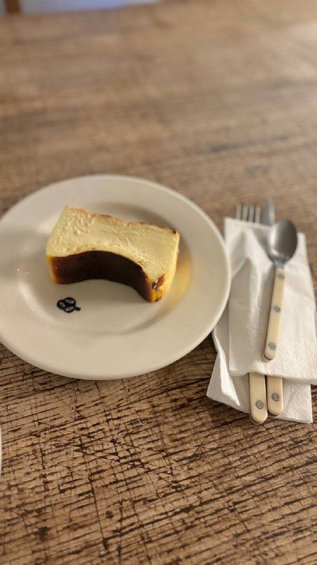 Кусок чизкейка на белой тарелке на грубой деревянной поверхности, рядом салфетки и приборы — боковой план, подчёркнута нежная текстура десерта.