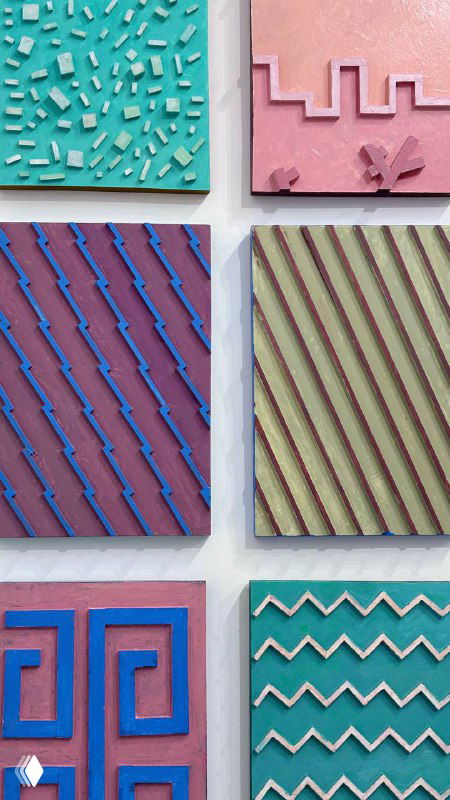 Ряды небольших цветных квадратных панно с геометрическими узорами и пастельными оттенками на белой стене галереи, демонстрация абстрактной серии работ.