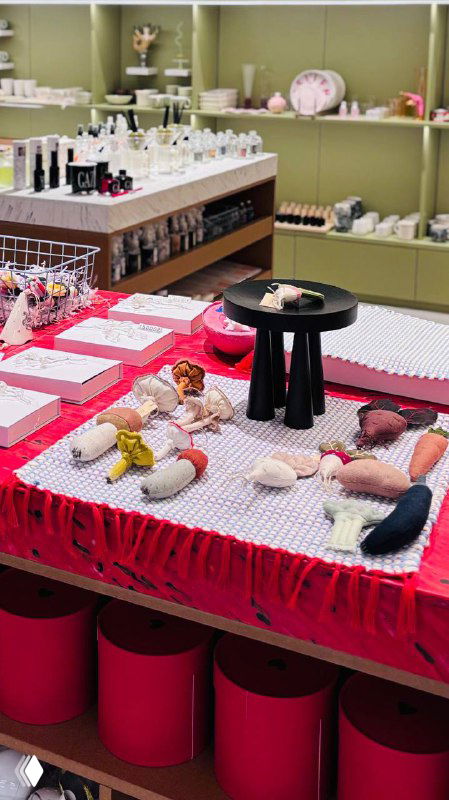 Ярко оформленный стол с текстильными изделиями и мелкими игрушками: на переднем плане мягкие куклы и элементы витрины магазина.
