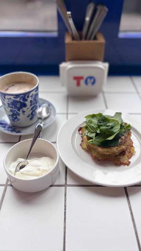 Тарелка с кабачковыми оладьями и зелёным салатом, миска сметаны и фарфоровая чашка с кофе на белой плитке