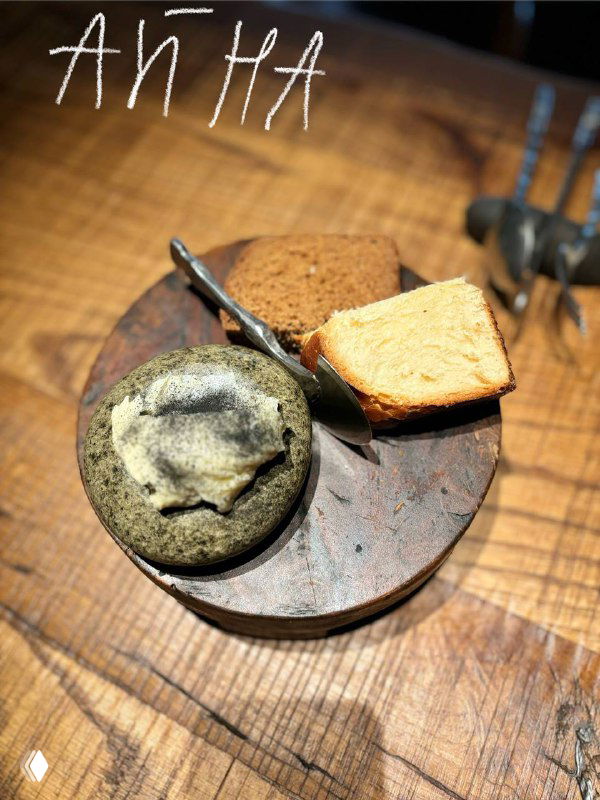 Две булочки на деревянной тарелке: молочный хлеб и бородинский с порцией домашнего масла, деревянный стол, надпись «АЙНА».