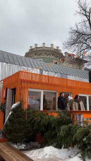 Оранжевый павильон или контейнер у входа во двор: декоративные деревянные элементы, окна и хвойные растения, вид на крышу соседнего здания.