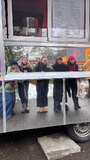 Группа людей у фудтрака с зеркальной поверхностью сервировочной стойки: несколько посетителей улыбаются, уличная обстановка и зимняя одежда.