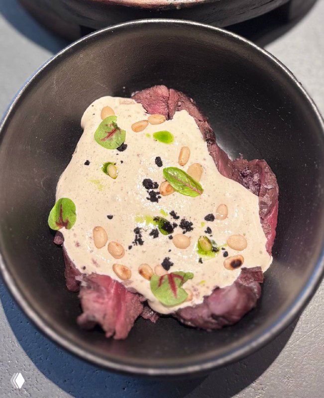 Крупный план блюда: тонко нарезанная оленина с муссом из ряпуши, кедровыми орешками и зелёными листиками, на чёрной тарелке.