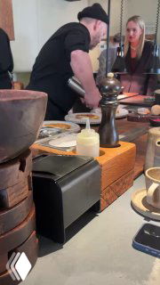 Шеф за барной стойкой в процессе готовки: посуда, соусы, мельница с перцем и наблюдающие гости, атмосферный кадр.