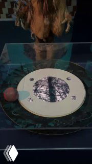 Экспонат с интерактивным столом и круговой панелью — часть инсталляции, рядом виднеется посетитель и тёмный интерьер зала.