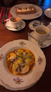 Тарелка с пельменями в соусе на столе, рядом чашки с чаем и кофе и десерт на заднем плане, тёплая ресторанная сервировка и свет.