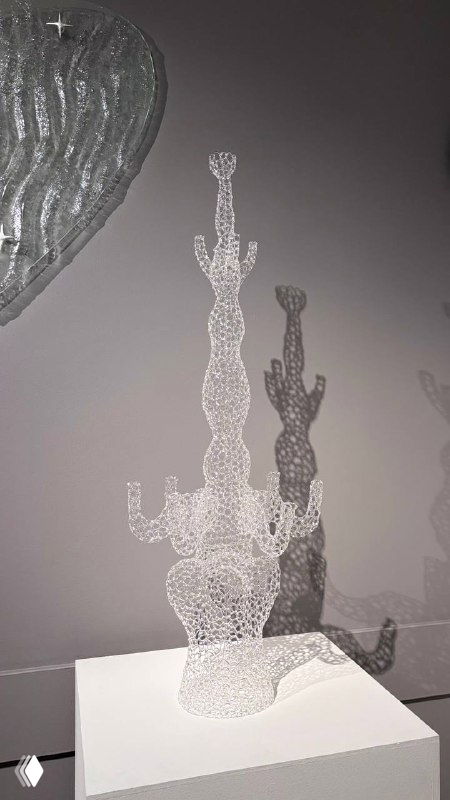 Прозрачная скульптура из точечных стеклянных элементов на пьедестале в галерее Heritage, отбрасывающая сложную тень на белую стену экспозиции.