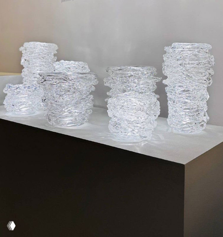 Ряд прозрачных стеклянных сосудов и форм, расположенных на постаменте в галерейном пространстве, акцент на преломлении света и текстуре стекла.