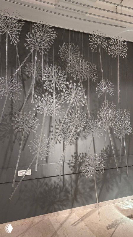 Стена, покрытая множеством тонких стеклянных элементов, напоминающих соцветия одуванчиков, которые создают сложные тени и ритм на задней стене выставки.