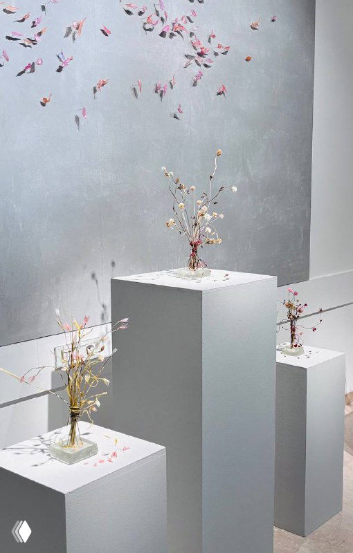 Три небольших композиции на постаментах с миниатюрными стеклянными букетами и разбросанными лепестками в минималистичном музейном интерьере Heritage.