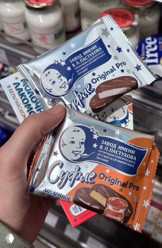 Рука держит упаковки суфле и чокопай завода им. В.П. Пастухова на полке магазина, видна бело‑синяя и серебристая упаковка с логотипом.