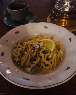 Тарелка пасты с ломтиком лимона, зеленью и кусочками морепродуктов; рядом чашка и стеклянный заварочный чайник на столе.