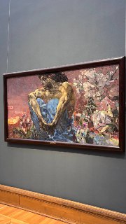 Фотография оформленной в раму картины на стене музея, вертикальный кадр, видна часть пола и нейтральный музейный интерьер.