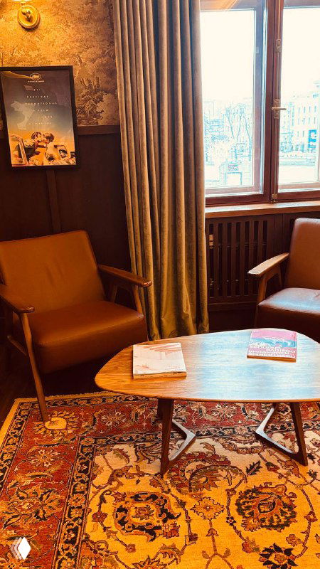 Небольшой зал ожидания с креслами у окна и журнальным столиком на ковровом покрытии, уютная зона перед началом показа в старом кинотеатре.