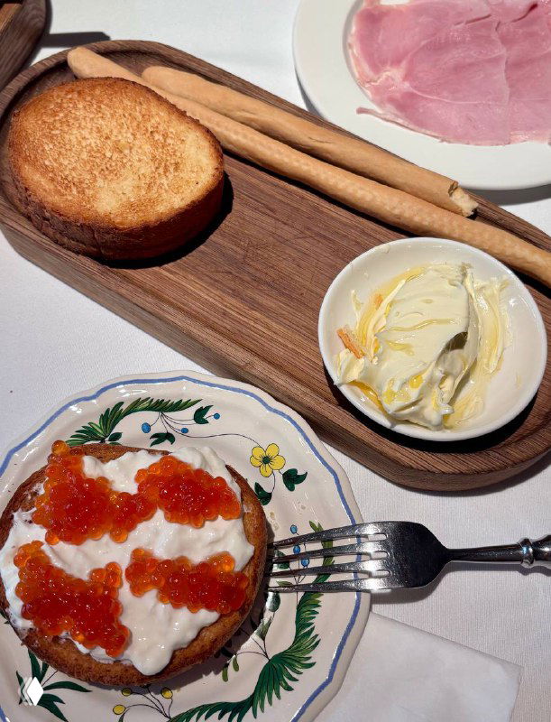 Крупный план: тёплая бриошь с нежной страчателлой и полосками красной икры на декоративной тарелке, рядом гриссини и масло с мёдом.