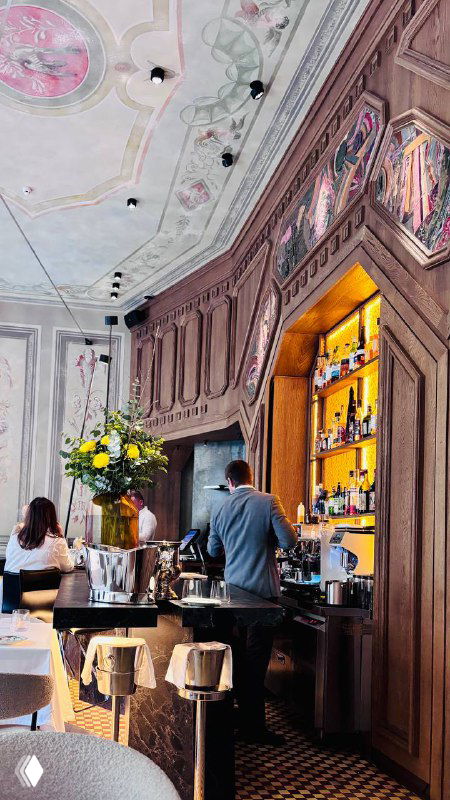 Интерьер ресторана Brera: барная зона с лепниной и витринами, цветы у стойки и посетители, заходящие в зал с высоким декоративным потолком.