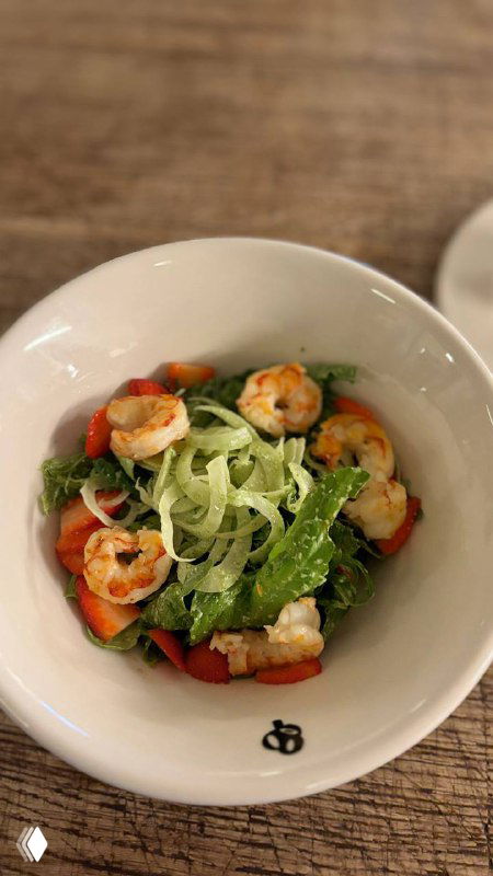 Тарелка салата с креветками, клубникой и фенхелем на столе в пекарне, видно свежие листья и яркую заправку, интерьер кафе на заднем фоне.