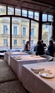 Вид из зала через большое окно на фасад соседнего здания и обедающих гостей у оконного ряда