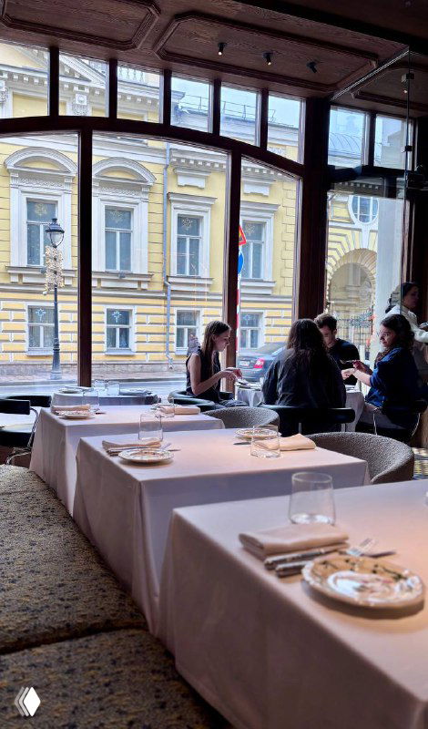 Вид через окно на фасад соседнего здания с классической архитектурой; посетители у оконных столиков завтракают в Brera на Большой Никитской.