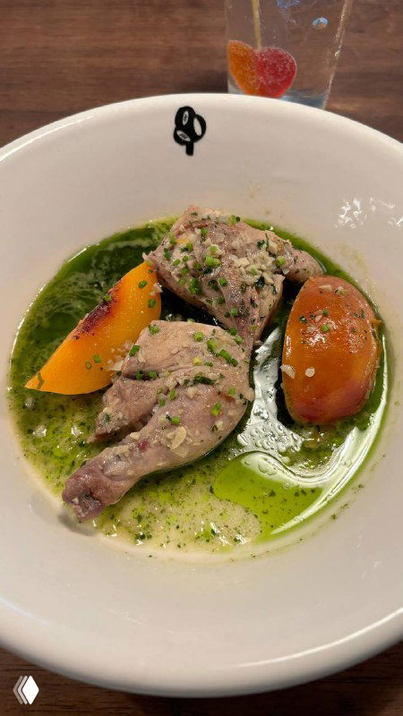 Блюдо с томлёным мясом птицы в белом вине, рядом дольки нектарина и зелёный масляный соус, подано в белой глубокой тарелке на столе.