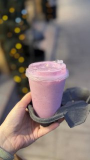 Рука держит розовый вишнёвый милкшейк в пластиковом стакане с крышкой, на размытом городском фоне видны огни и вечерняя сцена.