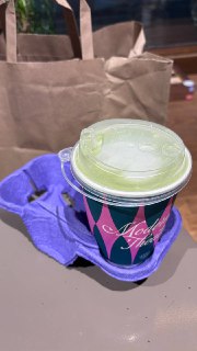 Бумажный стаканчик сети кофе с цветной подставкой стоит на столе рядом с бумажным пакетом и фиолетовой тканью в интерьере кафе.