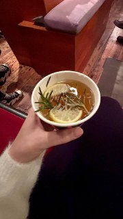 Крупный план бумажной чашки с тёплым чаем, в чашке видна долька лимона и веточка розмарина, рука держит чашку в уютном интерьере.