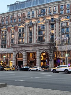 Фасад отеля Карлтон и вход ресторана Sartoria Lamberti на Тверской улице вечером, классическая архитектура, машины и прохожие у дороги.