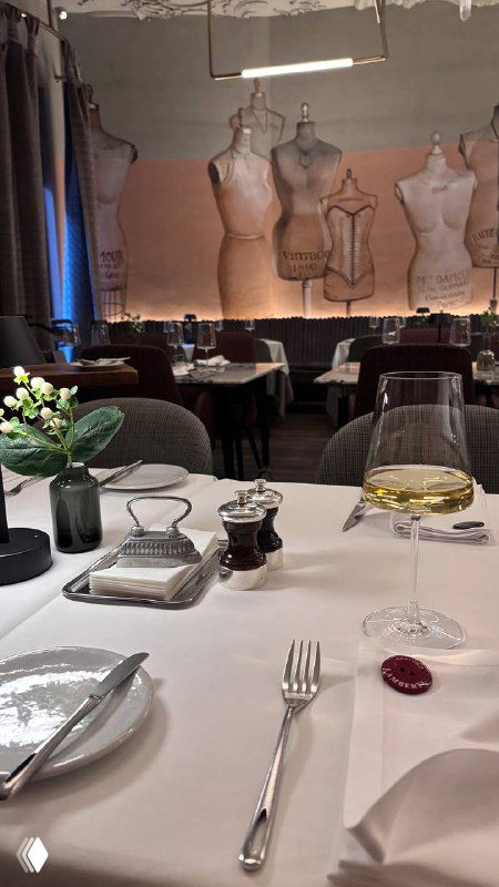 Крупный план сервировки за столом в Sartoria Lamberti: бокал вина, приборы и салфетка на фоне декоративных манекенов и мягкой подсветки интерьера.